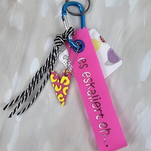 Könnte beinhalten: Ein pinkfarbener Schlüsselanhänger mit dem silbernen Schriftzug "es eskaliert eh...". Er hat einen blauen Karabinerhaken, einen herzförmigen Anhänger mit gelb-rosa Muster und eine schwarz-weiße Quaste.