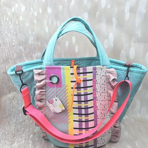 Könnte beinhalten: Eine türkisfarbene Tragetasche mit einer gemusterten Vorderseite aus Karos, gelben Streifen und einem Streusel-Druck. Die Tasche verfügt über rosa Rüschen, einen verstellbaren neongrünen Schulterriemen, zwei Tragegriffe und eine silberne Metallöse.