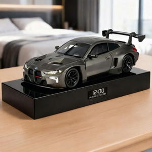 Op de afbeelding: Een grijs modelauto met zwarte wielen en een achterspoiler staat op een zwarte rechthoekige basis. De basis toont de tijd als 12:00 en de alarmstatus. De auto heeft een gedetailleerd ontwerp, inclusief koplampen en een rode accent op de voorkant.