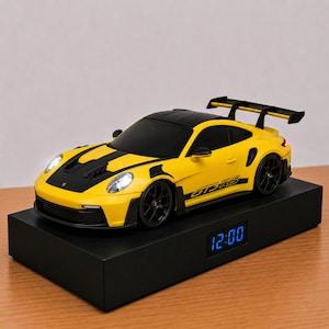 May include: A yellow and black Porsche GT3 RS model car with a large rear spoiler and illuminated headlights, mounted on a rectangular black base featuring a digital clock display that shows the time as 12:00 in bright blue glowing numbers.