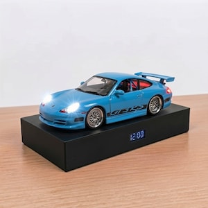 May include: A light blue Porsche 911 GT3 RS model car with illuminated white headlights and red taillights, mounted on a rectangular black base featuring a digital clock display showing the time 12:00 in blue LED numbers on the front side.