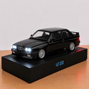 Puede incluir: Un coche a escala BMW M3 negro con faros blancos y luces traseras rojas iluminadas, montado sobre una base rectangular negra que cuenta con un reloj digital que muestra la hora 12:00 en una luz azul brillante, capturando el diseño clásico.