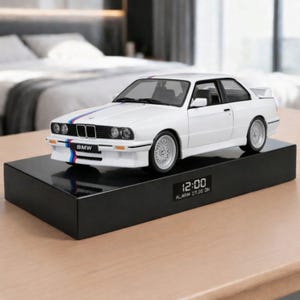Puede incluir: Un coche modelo BMW blanco con rayas de carreras azules y rojas se encuentra sobre una base rectangular negra. La base muestra la hora, 12:00, y las palabras "ALARM 07.00 ON". El coche tiene ruedas plateadas y el logotipo de BMW en la parte delantera.