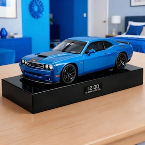 May include: A blue Dodge Challenger model car with black racing stripes sits atop a black rectangular base. The base features a digital clock displaying 12:00 and alarm settings. The car has black wheels and a sleek design.