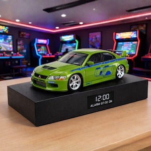 Peut inclure: Une voiture miniature verte et bleue avec des jantes argentées est posée sur un socle rectangulaire noir. La voiture a des rayures bleues et un aileron. L'heure affichée est 12h00, l'alarme est réglée à 07h00.