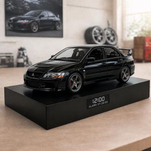 Peut inclure: Une voiture miniature noire avec des jantes argentées est posée sur un socle rectangulaire noir. La voiture est une berline avec un aileron arrière. Le socle affiche l'heure "12:00" et "ALARM 07:00 ON". Une photo encadrée d'une voiture similaire est en arrière-plan.