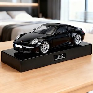 Op de afbeelding: Een zwart sportwagenmodel op een zwarte rechthoekige basis. De basis heeft een digitale klok met 12:00 en een alarm ingesteld op 07:00. De auto heeft zilveren wielen en een gestroomlijnd ontwerp.