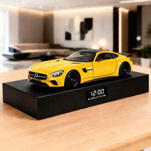 May include: A yellow sports car model with black accents and wheels sits atop a black rectangular base. The base displays a digital clock showing 12:00 and alarm settings. The car model is a detailed replica.