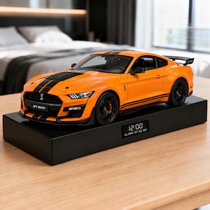Könnte beinhalten: Ein orangefarbenes Ford Mustang GT500 Modellauto mit schwarzen Rennstreifen und schwarzen Rädern, montiert auf einem rechteckigen schwarzen Sockel. Der Sockel verfügt über eine Digitalanzeige mit 12:00 und ALARM 07:00 ON. GT500 Emblem am Kühlergrill.