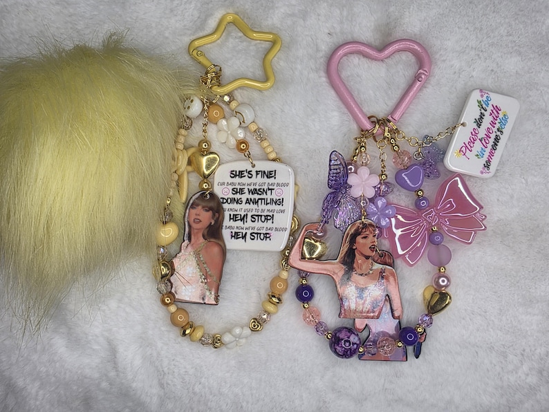May include: Two keychains with celebrity images and beaded accents. One has a yellow star-shaped clip and a yellow pom-pom. The other has a pink heart-shaped clip and a pink bow. Both have text that reads "SHE'S FINE!" and "Please don't be in love with someone else."