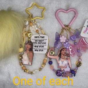 May include: Two keychains with beaded accents and star and heart-shaped clasps. One features a portrait and text that reads "SHE'S FINE!" The other has a portrait and a pink bow. A yellow pom-pom is on the left.