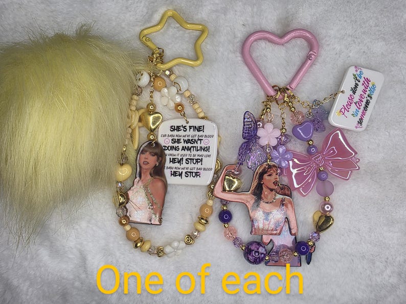 May include: Two keychains with beaded accents and star and heart-shaped clasps. One features a portrait and text that reads "SHE'S FINE!" The other has a portrait and a pink bow. A yellow pom-pom is on the left.