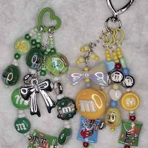 Puede incluir: Dos llaveros de cuentas con dijes temáticos de dulces. Un llavero presenta cuentas verdes y plateadas con dijes de M&M's verdes y un lazo plateado. El otro tiene cuentas amarillas, azules y rojas con dijes de M&M's y un cierre en forma de corazón.