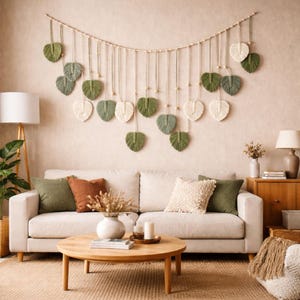 May include: A macramé wall hanging featuring leaf-shaped designs in green and cream hues. The leaves are suspended from a wooden dowel with wooden beads. It hangs above a cream sofa with decorative cushions, creating a cozy atmosphere.