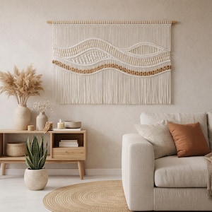 Può includere: Un grande arazzo da parete in macramè color crema con un design a onde, con dettagli intrecciati e annodati e un bordo inferiore con frange. L'arredamento della parete è montato su un'asta di legno. La stanza comprende anche un divano beige, un mobile in legno e un tappeto rotondo in juta.