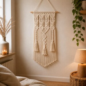 Puede incluir: Un tapiz de macramé color crema suspendido de un listón de madera horizontal. La pieza presenta intrincados patrones de nudos, incluyendo columnas verticales, formas de diamante y tres borlas decorativas en el borde inferior.