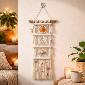 May include: A cream-colored macrame wall hanging with a wooden dowel and seashells. The design features a central orange seashell, a starfish, and various other shells. The piece is suspended from a wall with a decorative hook, creating a coastal theme.