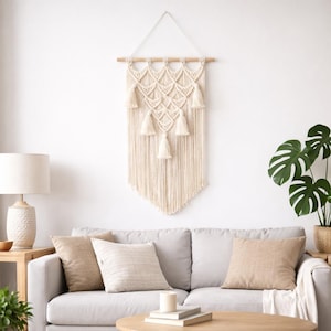 May include: Cream macrame wall hanging with a geometric pattern and tassels, suspended from a wooden dowel. The wall art is displayed against a white wall. The decor is part of a living room setting with a sofa, cushions, and a lamp.