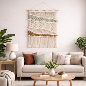 May include: A cream-coloured macrame wall hanging with a wooden dowel. The design features layers of textured weaving in cream, sage green, and brown. The wall hanging is displayed above a cream-coloured sofa with decorative cushions.