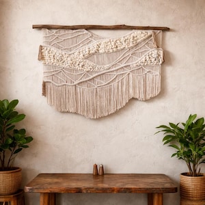 May include: A beige macrame wall hanging suspended from a natural wooden branch. The wall art features woven and knotted patterns, with a fringed bottom. The neutral colour palette and organic textures create a bohemian aesthetic, suitable for home decor.