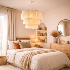 Könnte beinhalten: Ein Schlafzimmer mit einem cremefarbenen, dreistufigen Kronleuchter über einem Bett mit weißer Bettwäsche und Kissen. Eine Holzkommode, ein runder Spiegel und ein Weidenkorb tragen zum neutralen Farbschema des Raumes bei.