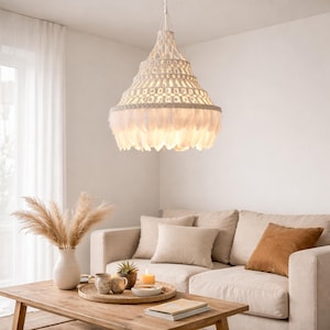 Könnte beinhalten: Eine weiße Makramee-Hängeleuchte mit abgestuftem Design und einem Saum aus weißen Federn. Die Leuchte hängt über einem Wohnzimmer mit beigem Sofa, Holztisch und dekorativen Gegenständen.