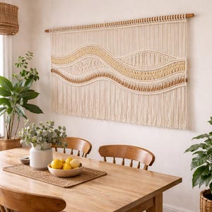 May include: A cream-coloured macrame wall hanging with a wavy design in shades of beige and brown. The textile art piece is mounted on a wooden rod and hangs above a wooden dining table. The room is decorated with plants and a bowl of lemons.