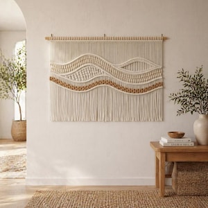 Puede incluir: Un gran tapiz de macramé color crema con un diseño ondulado. La pieza presenta una varilla de madera en la parte superior y flecos en la parte inferior. Los detalles tejidos incluyen secciones de color beige y crema. La decoración de la pared se exhibe en una habitación de tonos neutros.