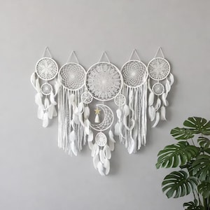 May include: A set of white dream catchers with detailed web patterns and dangling feathers. The dream catchers are of different sizes and shapes, with a crescent moon and star feature on the central piece. The wall decor is set against a grey backdrop.