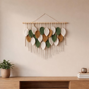 May include: A wall hanging featuring a wooden dowel with a series of white, green, and gold leaf-shaped macrame elements. The leaves are arranged in a cascading pattern, with long white fringe hanging below. The wall decor is displayed against a neutral wall.