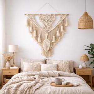 Op de afbeelding: Een slaapkamer met een crèmekleurige macramé wandhanger boven een bed met neutrale beddengoed. De macramé heeft kwasten en een bladvorm. Houten nachtkastjes met lampen en een geweven hanglamp maken de gezellige, bohemien sfeer compleet.