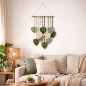Pode incluir: Uma tapeçaria de parede em macramé com folhas em forma de coração em tons de verde e creme, suspensa de um pino de madeira. As folhas estão dispostas em um padrão em cascata, criando uma estética natural e texturizada. Esta peça decorativa adiciona um toque boêmio a qualquer espaço interior.