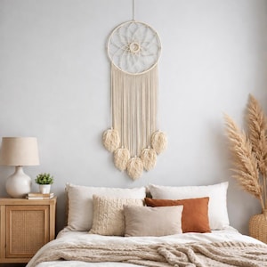 May include: A cream-coloured macrame dream catcher suspended on a light grey wall. The dream catcher features a circular woven design with long, dangling strands and leaf-shaped accents. The bedroom setting includes a bed with pillows and a side table.