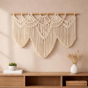 May include: Cream-coloured macrame wall hanging with a geometric design, suspended from a natural wooden dowel. The piece features intricate knotting patterns and long fringe, adding a bohemian touch to home decor. The wall hanging is displayed against a neutral-toned wall.
