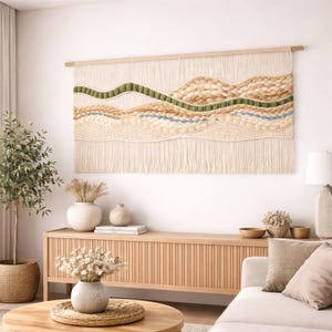 May include: A large, textured wall hanging with a landscape design. The piece features cream, tan, green, and blue yarn, creating a layered, organic pattern. It is mounted on a natural wood dowel, adding a touch of warmth to the neutral color palette.