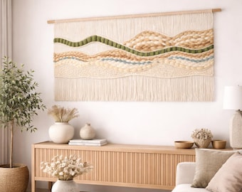 May include: A large, textured wall hanging with a landscape design. The piece features cream, tan, green, and blue yarn, creating a layered, organic pattern. It is mounted on a natural wood dowel, adding a touch of warmth to the neutral color palette.