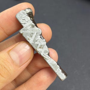 Pode incluir: Um fragmento de meteorito metálico alongado com um padrão de Widmanstätten distinto de linhas cristalinas cruzadas. A superfície apresenta um acabamento polido cinza-prateado com bordas escuras e irregulares e um perfil irregular.