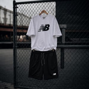 May include: A white short-sleeved t-shirt featuring a black New Balance logo on the chest, paired with black athletic shorts that have a white drawstring and a small white New Balance logo on the lower left leg, displayed on a wooden hanger.