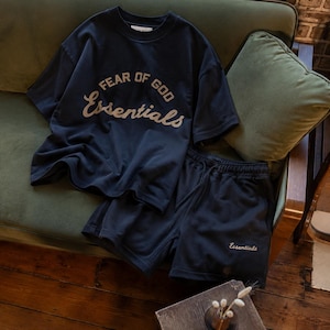 May include: A dark navy blue short-sleeve t-shirt and matching shorts set. The t-shirt features the text FEAR OF GOD Essentials in a light-colored font across the chest. The shorts have the word Essentials printed in a light font on the lower leg.