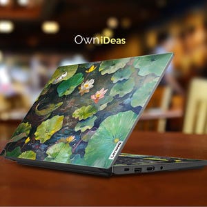 Puede incluir: Un ordenador port&aacute;til con un dise&ntilde;o floral colorido en la tapa. El dise&ntilde;o presenta nen&uacute;fares y hojas en tonos de verde, amarillo y rosa. El port&aacute;til est&aacute; cerrado y el logotipo de Lenovo es visible en la esquina inferior derecha de la tapa.