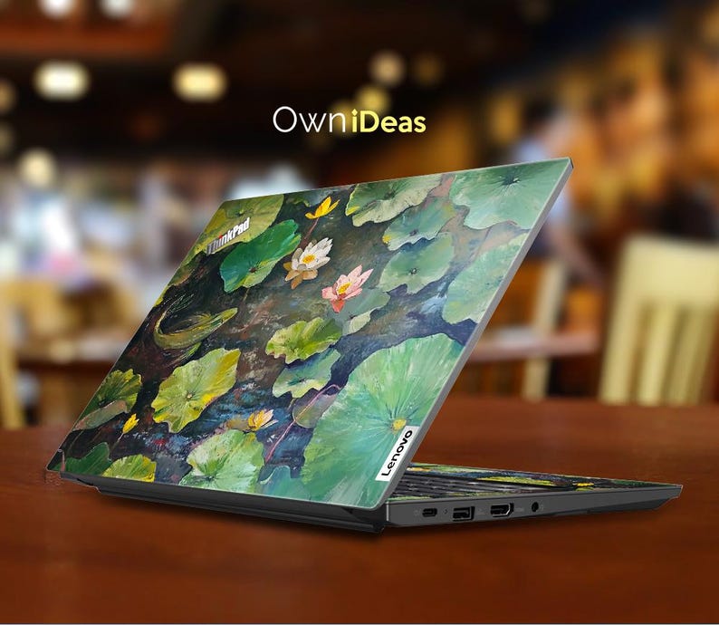 Puede incluir: Un ordenador port&aacute;til con un dise&ntilde;o floral colorido en la tapa. El dise&ntilde;o presenta nen&uacute;fares y hojas en tonos de verde, amarillo y rosa. El port&aacute;til est&aacute; cerrado y el logotipo de Lenovo es visible en la esquina inferior derecha de la tapa.