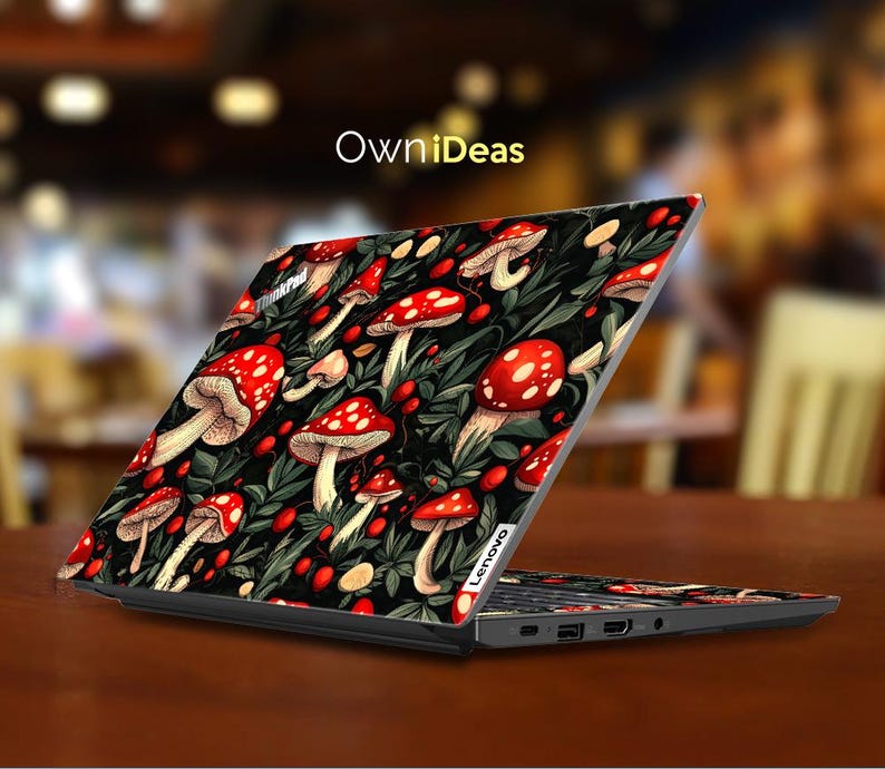 May include: A Lenovo laptop with a custom skin featuring a pattern of red mushrooms with white spots and green foliage on a black background. The laptop is open on a brown surface, with the text "OwniDeas" visible.