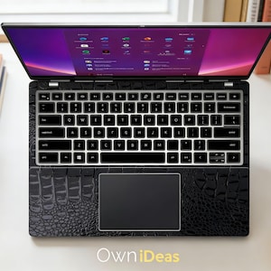 May include: Black laptop with a textured, crocodile-like pattern. The keyboard has white keys with black lettering. The screen displays a colorful interface. The laptop is open on a white surface, with books and a coffee cup nearby. The text "Own iDeas" is visible.