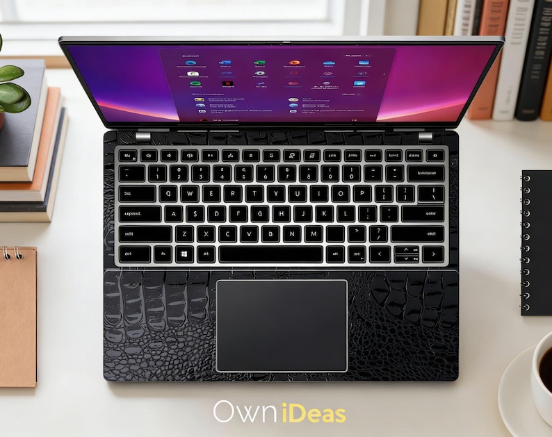 May include: Black laptop with a textured, crocodile-like pattern. The keyboard has white keys with black lettering. The screen displays a colorful interface. The laptop is open on a white surface, with books and a coffee cup nearby. The text "Own iDeas" is visible.