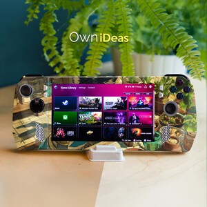 May include: A handheld games console with a custom skin featuring a botanical design. The screen displays a game library with various game titles. The console is resting on a white stand. The text "OwniDeas" is visible.