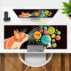以下が含まれることがあります： カラフルな惑星を吹き出す漫画の猫が描かれた長方形のマウスパッド。惑星は様々なサイズと色で、黒い星空の背景に配置されています。「OwniDeas」の文字が中央にあります。