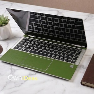 May include: A laptop with a green skin on a white marble surface. The laptop is open, displaying the keyboard and screen. A small succulent plant in a white geometric pot, a brown leather notebook, a gold pen, and a coaster are also on the surface.
