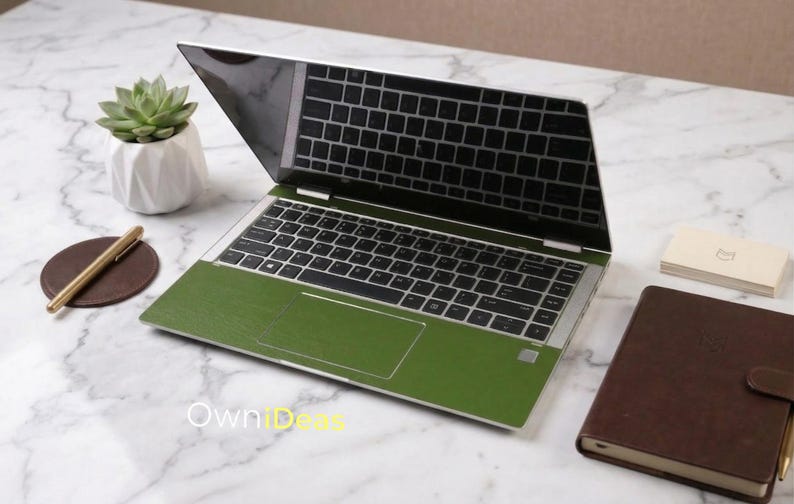 May include: A laptop with a green skin on a white marble surface. The laptop is open, displaying the keyboard and screen. A small succulent plant in a white geometric pot, a brown leather notebook, a gold pen, and a coaster are also on the surface.