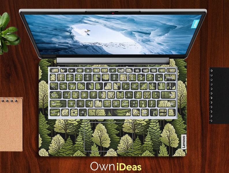 May include: A laptop computer with a green and white forest design on the keyboard and trackpad. The laptop is closed and the screen is showing a blue and white image of a snowy mountain.