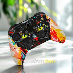 May include: Xbox controller with a black cat design and yellow eyes, set against colourful paint splatter. The controller has black buttons and joysticks, with the Xbox logo. The design includes the text "OwniDeas".